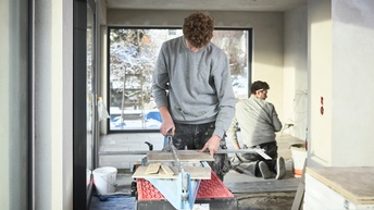 Ein junger Mann schneidet Fliesen - im Hintergrund werden sie von seinem Kollegen verlegt. 