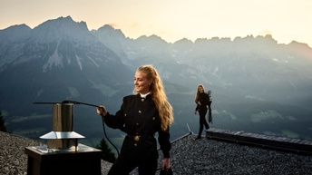Eine Person auf einem mit Kies ausgelegten Flachdach am Kamin. Im Hintergrund eine Kollegin und eine sonnige Alpenkulisse