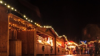 Mehrere Holzhütten stehen auf einem asphaltierten Weg. Die Hütten sind vorne teilweise offen. Darin befinden sich verschiedene Objekte. Auf den Dächern sind Lichterketten. Im Hintergrund sind viele Menschen. Der Himmel ist sehr dunkel