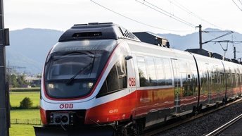 Zug fährt auf Gleisen daneben zeigt sich eine ländliche Kulisse und im Hintergrund grüne, Berge mit Forstbeständen