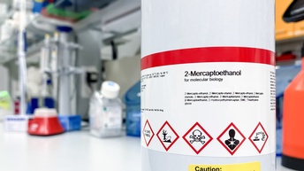 Weißer Metalleimer mit rotweißem Etikett mit schwarzer Aufschrift 2-Mercaptoethanol und verschiedenen weißen Warnzeichen mit roter Umrandung wie Totenkopf, darunter weiteres gelbes Etikett mit Aufschrift Vorsicht, Überdruck möglich