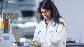 Person in weißem Arbeitskittel und Gummihandschuhe mit Brille blickt auf Gerät