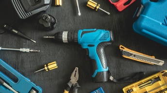 Auf dunklem Untergrund liegen nebeneinander verschiedene Werkzeuge, wie ein Hammer, ein Stanleymesser, Zangen, Schraubenzieher, Bohrer, Aufsätze zum Bohren und Schrauben.