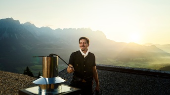 Ein junger Mann auf einem mit Kies ausgelegten Flachdach am Kamin. Im Hintergrund eine sonnige Alpenkulisse