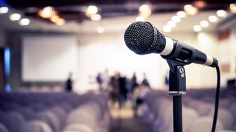 Nahaufnahme eines Mikrofons vor einem Raum mit Sesselreihen und ein paar stehenden Personen in der Unschärfe