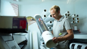 Ein junger Mann hält eine bedruckte Rolle Papier. Im Hintergrund Folien- und Papierrollen