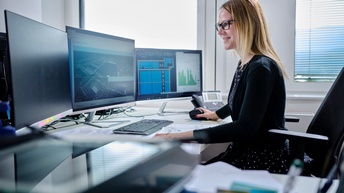 Eine junge Frau sitzt im Büro vor drei Bildschirmen und plant eine große Photovoltaik-Anlage