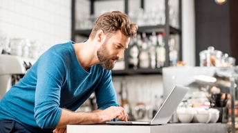 Person mit Bart in blauem Pullover über Laptop gebeugt, im Hintergrund Utensilien einer Bar