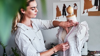 Eine Person mit hellen Haaren steht seitlich neben einer Modepuppe, auf der ein gestreiftes Oberteil hängt. Die Person hält mit beiden Händen ein Maßband von der linken Schulter der Puppe über die Brust und blickt darauf