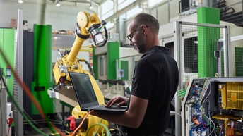 Ein Mann arbeitet stehend an seinem Laptop, im Hintergrund ein Robotergreifarm, den er einstellt