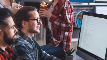 Seitliche Aufnahme zweier neben einem Bildschirm sitzender Personen. Daneben sind zwei weitere stehende Personen. Alle blicken auf den Bildschirm, auf dem mehrere Zeilen sind