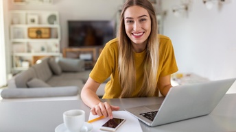 Porträt einer Person mit langen, hellen Haaren, die an einem Tisch in einem Wohnessbereich sitzt. Vor der Person sind ein aufgeklappter Laptop, ein Zettel, ein Handy, ein Stift und eine Tasse