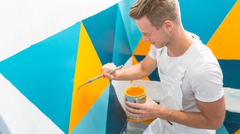Person in weißem Arbeitsgewand streicht gelbe Farbe an Wand, die mit blauen, gelben und türkisen Dreiecken gemustert ist