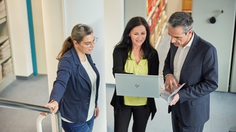 Drei Personen stehen in einem Flur, eine Person in der Mitte hält einen Laptop in Händen, eine Person neben ihr blickt gespannt darauf, während ein geöffnetes Buch in Händen gehalten wird