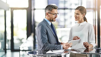 Zwei Personen stehen an Tischen in Unterhaltung, eine Person hält Tablet in Händen, am Tisch Dokument mit Stift liegend