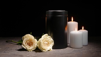 Schwarze Urne, nebenliegend zwei weiße Rosen und drei brennende weiße Kerzen, Hintergrund schwarz