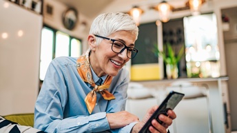 Person mit kurzen weißblonden Haaren und Brille sitzt in einem Wohnraum und blickt freudig auf ein Smartphone