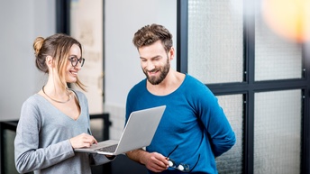 Zwei lächelnde Personen auf aufgeklapptes Notebook blickend