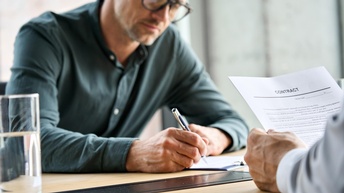 Person mit Brillen unterfertigt Dokument, Ausschnitt anderer Person, die Dokument mit Schriftzug Contract in Händen hält 