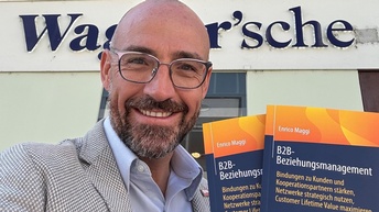Ein lächelnder Mann mit Brille hält mehrere Exemplare eines Buches in den Händen und steht vor einer Buchhandlung.