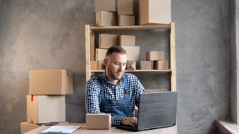 Eine Person sitzt an einem Holztisch. Vor ihr ist ein aufgeklappter Laptop, auf den sie blickt, sowie ein kleines Päckchen und ein Klemmbrett. Hinter der Person stehen weitere Kartons am Boden und in einem Regal