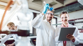 Drei Personen in weißen Kitteln mit Schutzbrillen in Labor, eine Person schwenkt Glas mit Flüssigkeit, eine andere blickt auf Clipboard