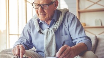 Person mit weißem Haar und Brille bedient mit einer Hand Taschenrechner und notiert mit zweiter Hand in Buch