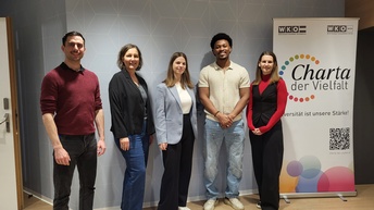 Fünf Personen stehen nebeneinander vor einer grauen Wand. Neben ihnen steht ein Banner, auf dem unter anderem Charta der Vielfalt steht