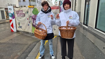 Porträt von zwei Personen, die auf einem Gehweg mit jeweils einem Einkaufskorb stehen. Im Einkaufskorb sind klein verpackte Schokokugeln. Beide Personen tragen weiße Jacken, auf denen mein Kaufstadt Wien steht