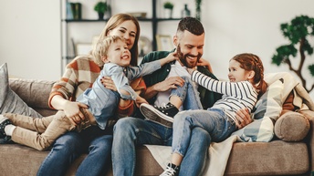 Zwei Erwachsene und zwei Kinder sitzen auf einem hellbraunen Sofa. Die Kinder liegen auf den Erwachsenen. Alle interagieren miteinander. 