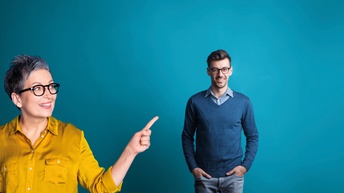 Vor blauem Hintergrund steht rechts eine Person. Links im Bild davor steht eine weitere Person, die auf die andere Person zeigt