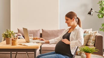 Schwangere Person sitzt freudig bei einem Wohnzimmertisch und arbeitet mit einem Laptop während im Hintergrund eine Couch sowie Pflanzen stehen
