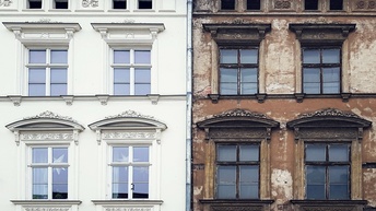 Mauer an Mauer stehen nebeneinander zwei mehrstöckige Häuser. Das linke Haus hat eine weiße, renovierte Fassade. Die Fassade des rechten Hauses ist bräunlich und blättert an vielen Stellen ab