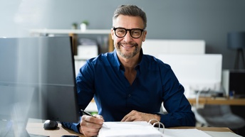 Porträt einer an einem Tisch sitzenden Person mit kurzen, grauen Haaren, Bart und Brille. Direkt vor der Person ist ein aufgeklappter mit einem Stapel Papier. Die Person hält einen Stift. Daneben ist ein Computer