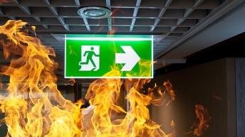 Ein grünes Schild hängt von der Decke. Auf dem Schild ist eine Silhouette einer Person, die durch eine Tür läuft. Daneben zeigt ein Pfeil nach rechts. Unter und links neben dem Schild ist eine rote Flamme.