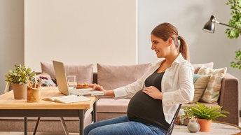 Schwangere Person sitzt freudig bei einem Wohnzimmertisch und arbeitet mit einem Laptop während im Hintergrund eine Couch sowie Pflanzen stehen