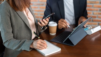 Zwei Personen stehen an einem Tisch. Die linke Person notiert etwas auf einem Notizbuch. Zwischen den Personen steht ein Tablet