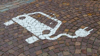 Auf einem Kopfsteinpflaster sieht man den Hinweis auf eine E-Lade-Station für ein Elektroauto als Bodenmarkierung in weißer Farbe. Das Symbol dominiert das Bild, das Kopfsteinpflaster ist nass vom Regenwetter. 