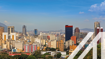 Stadtansicht von Caracas: Skyline mit zahlreichen Wolkenkratzern in Downtown Caracas, Hauptstadt von Venezuela, unter blauem Himmel, gelegen vor einer Bergkette. Über das Bild wurde ein weißes Austria A gelegt.