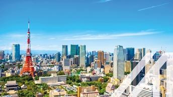 Stadtansicht von Tokio mit vielen Hochhäusern und dem bekannten rot-weißen Tokyo Tower unter blauem Himmel. Über das Bild wurde ein weißes Austria A gelegt.