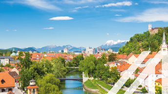 Blick auf Sloweniens Hauptstadt Laibach mit vielem weißen Häusern und roten Dächern. In der Mitte schlängelt sich ein Fluss mit Brücken durch die Stadt, rechts im Bild sieht man auf einer Anhöhe eine Burg, im Hintergrund verschneite Berggipfel. 