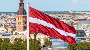 Die Flagge Lettlands weht im Wind unter blauem Himmel vor einer Stadtansicht von Riga.