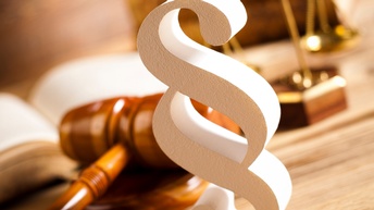 Nahaufnahme eines Paragraphensymbols, das auf einer hölzernen Oberfläche steht. Dahinter liegen ein Hammer aus Holz, ein aufgeklapptes Buch und eine Waage. Die Objekte befinden sich in einem hell ausgelichteten Raum