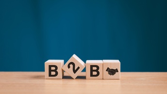 Auf einem Tisch aus hellem Holz sind vier Würfel platziert, wobei der zweite schief liegt, die anderen sind aufgestellt. Auf den Würfel steht B2B und am vierten Würfel ist das Symbol von sich schüttelnden Händen. Der Hintergrund ist blau