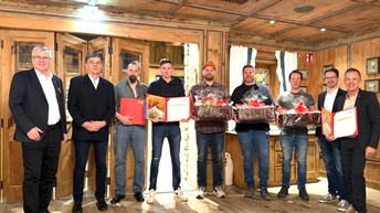 Mehrere Personen stehen nebeneinander in einem holzgetäfelten Innenraum. Einige halten geöffnete Urkunden oder Geschenkkörbe in den Händen. Alle blicken in die Kamera und stehen in einer Reihe vor einer geöffneten Tür.