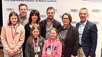 Eine Gruppe von acht Personen steht vor einem Hintergrund mit dem Logo der Wirtschaftskammer Tirol. Vorne stehen drei Kinder mit Medaillen um den Hals; zwei tragen Nike-Pullover. Hinter ihnen stehen fünf Erwachsene – drei Männer und zwei Frauen – in festlicher, aber alltäglicher Kleidung. Alle blicken lächelnd in die Kamera. Die Atmosphäre wirkt wie eine Preisverleihung oder Ehrung im Bezirksverband Landeck.