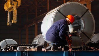 Person mit orangem Helm hockt in Rückenansicht vor großer Stahlspule in Halle und befestigt Kette, im Hintergrund gelbes Hebeelement von der Decke hängend