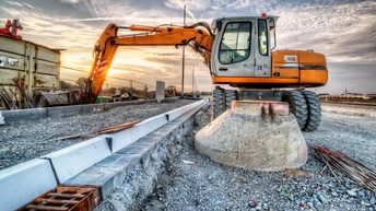 Ein Bagger steht auf einer Baustelle einer Straße. Links neben dem Bagger ist eine Erhöhung für einen Gehsteig. Neben dem Bagger liegen Metallstäbe