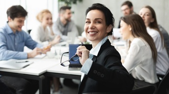 Person mit dunklen geschlossenen Haaren und Businesskleidung hält eine Brille in der Hand und blickt freudig in die Kamera, während im Hintergrund weitere Personen am selben Besprechungstisch sitzen