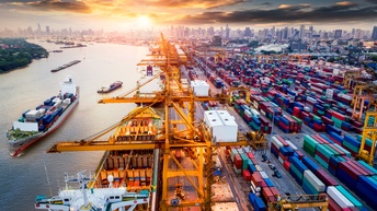 Top-Shot vieler bunter, übereinander und nebeneinander gestapelter Container. Links daneben ist ein Gewässer, auf dem unterschiedlich große Schiffe, beladen mit Container sind. Im Hintergrund sind viele hohe Wolkenkratzer.